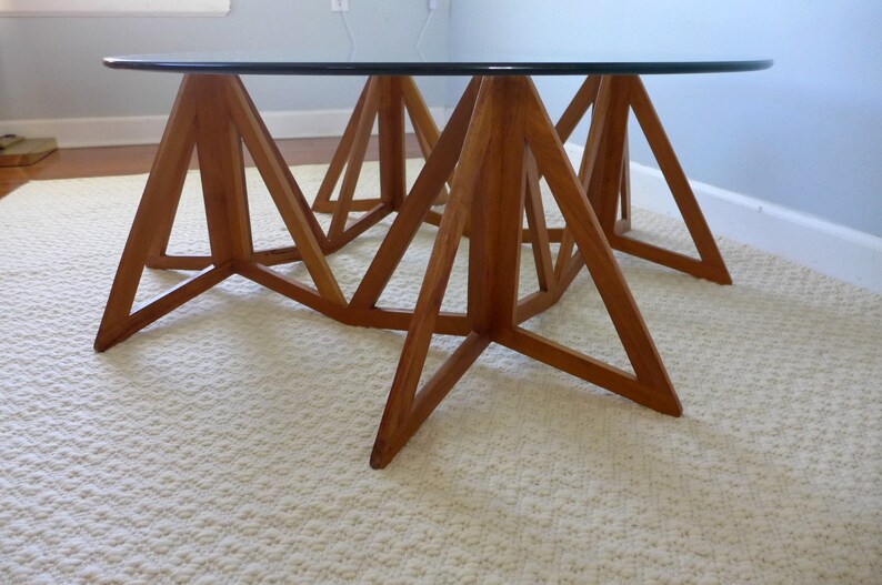 May include: A round glass top coffee table with a unique wooden base made of triangular shapes. The base is made of light brown wood and has a geometric design.