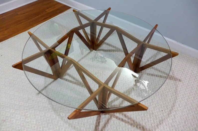 May include: A round glass-top coffee table with a unique geometric wooden base. The base is made of multiple pieces of wood that are joined together to create a starburst pattern.