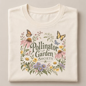 Könnte beinhalten: Ein cremefarbenes T-Shirt mit einem Grafikdruck aus verschiedenen Wildblumen, darunter rosa Sonnenhüte, weiße Gänseblümchen und violette Astern, die den Text "Pollinator Garden SOCIETY" umgeben, ergänzt durch orangefarbene und gelbe Schmetterlinge.