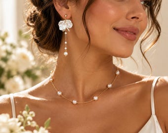 Op de afbeelding: Een goudkleurige ketting met gelijkmatig verdeelde witte parels aan een fijne ketting, gecombineerd met een bijpassende oorhanger met een wit bloemblaadjesvormig accent aan een dunne gouddraad met kleine pareldetails.