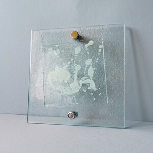 May include: Square glass decorative piece with a textured surface and a smaller square inset. The inset has a white, abstract design. Two metal fasteners secure the glass. A modern art piece.