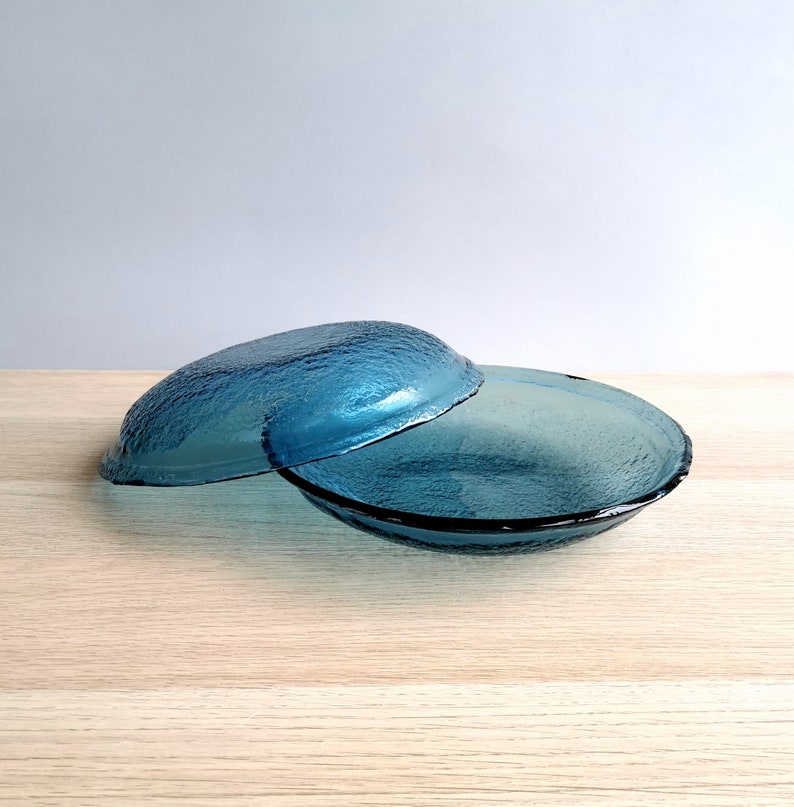 May include: Two blue glass bowls with a textured surface. One bowl is partially covering the other. The bowls are a vibrant shade of blue and appear to be made of glass. The bowls are sitting on a light-colored wooden surface.