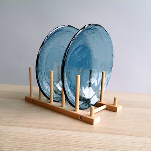 May include: Two blue glass plates with textured surfaces are displayed in a wooden plate rack. The plates have irregular edges and a light blue hue. The rack is made of light-colored wood.