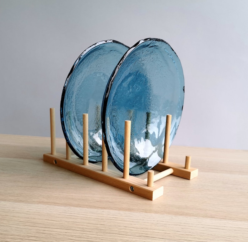 May include: Two blue glass plates with textured surfaces are displayed in a wooden plate rack. The plates have irregular edges and a light blue hue. The rack is made of light-colored wood.