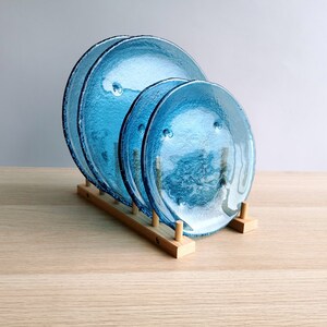 May include: Three textured, turquoise glass plates of varying sizes are displayed in a wooden plate rack. The plates have a bubbly texture and a dark blue rim. The wooden rack is made of light-colored wood.