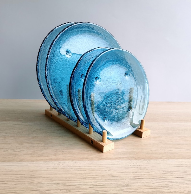May include: Three textured, turquoise glass plates of varying sizes are displayed in a wooden plate rack. The plates have a bubbly texture and a dark blue rim. The wooden rack is made of light-colored wood.