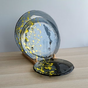 May include: Three black glass plates with yellow splatter paint. The plates are stacked on a wooden stand.
