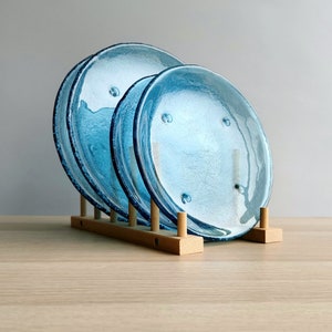 May include: A set of three blue glass plates with textured surfaces and dark blue rims, displayed in a wooden plate rack. The plates have a slightly irregular shape and are arranged on a light-colored wooden surface.