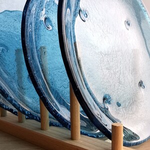 May include: A set of three blue glass plates with a wave-like design, displayed in a wooden plate rack. The plates have a textured surface and varying shades of blue, creating a water-like effect. The wooden rack has a natural finish.