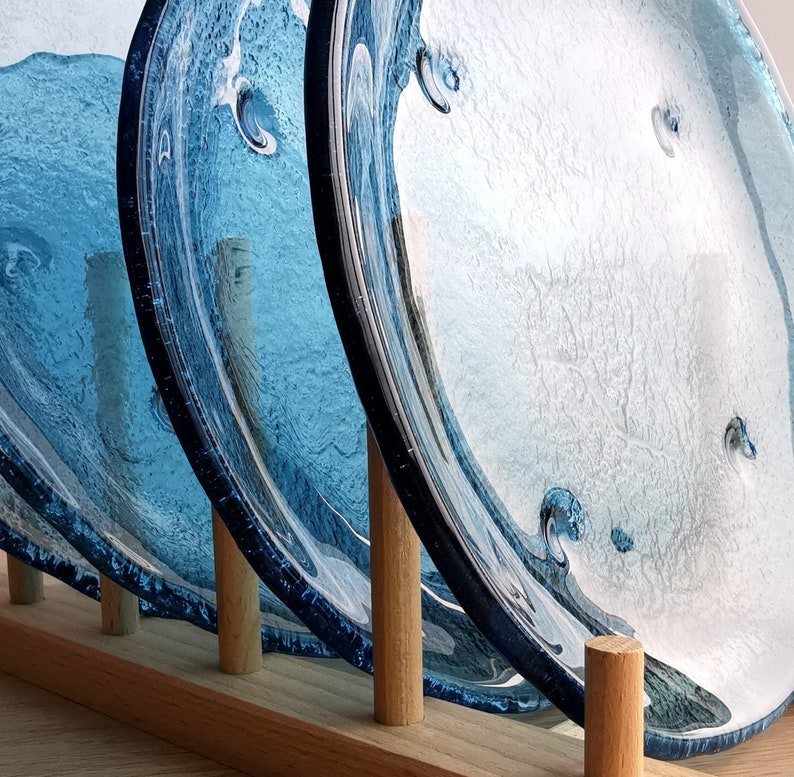 May include: A set of three blue glass plates with a wave-like design, displayed in a wooden plate rack. The plates have a textured surface and varying shades of blue, creating a water-like effect. The wooden rack has a natural finish.