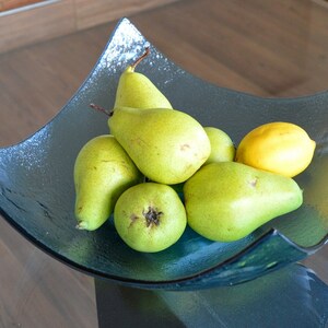 May include: A glass fruit bowl filled with green pears and a yellow lemon. The bowl has a unique, angular shape and a dark teal color. The pears are ripe and ready to eat, and the lemon adds a bright contrast.