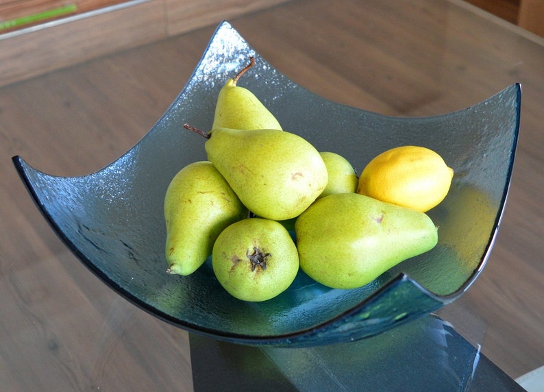 May include: A glass fruit bowl filled with green pears and a yellow lemon. The bowl has a unique, angular shape and a dark teal color. The pears are ripe and ready to eat, and the lemon adds a bright contrast.