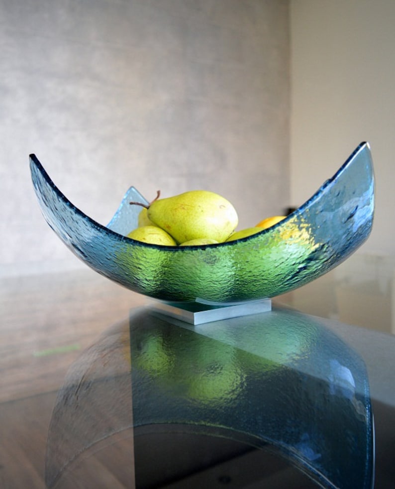May include: A textured, teal-colored glass bowl, filled with green pears. The bowl has a unique, curved shape and sits on a small, silver-colored base. The pears are ripe and ready to eat, adding a pop of color.