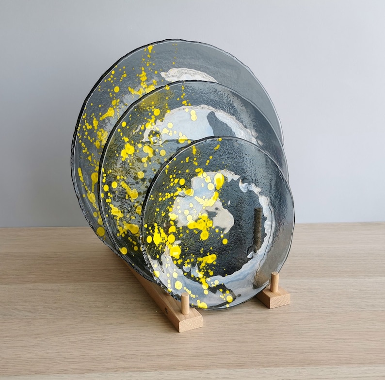 May include: Set of four glass plates with a black and yellow speckled design. The plates are stacked on a wooden stand.