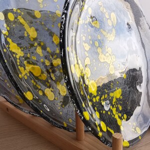 May include: Three glass plates with a black and white background and yellow splatter paint. The plates are stacked on a wooden stand.