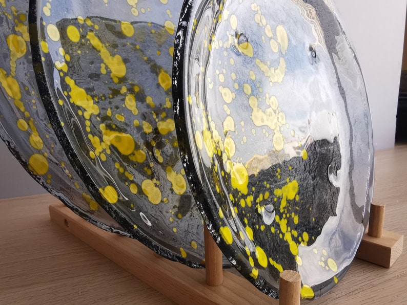 May include: Three glass plates with a black and white background and yellow splatter paint. The plates are stacked on a wooden stand.