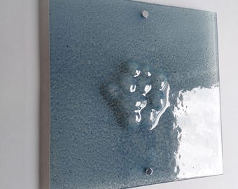 May include: Square, textured, blue glass wall art with a raised, abstract design. The artwork is mounted with two silver fasteners. The glass has a speckled appearance, and the raised design is a lighter shade of blue.