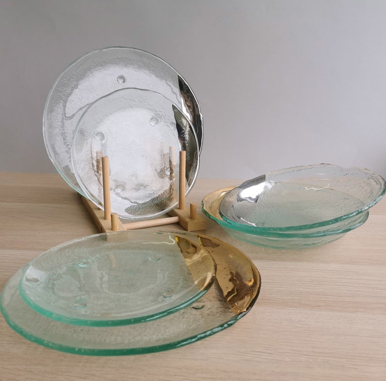 May include: Set of four clear glass plates with a textured surface. Two plates are stacked on a wooden stand, and two plates are stacked on a gold-rimmed plate.