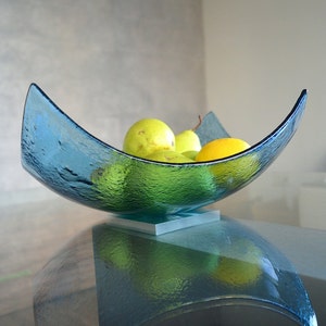 May include: A textured, turquoise glass bowl in a crescent shape, resting on a square metal base. The bowl is filled with green pears and a yellow lemon. The bowl is sitting on a glass table.