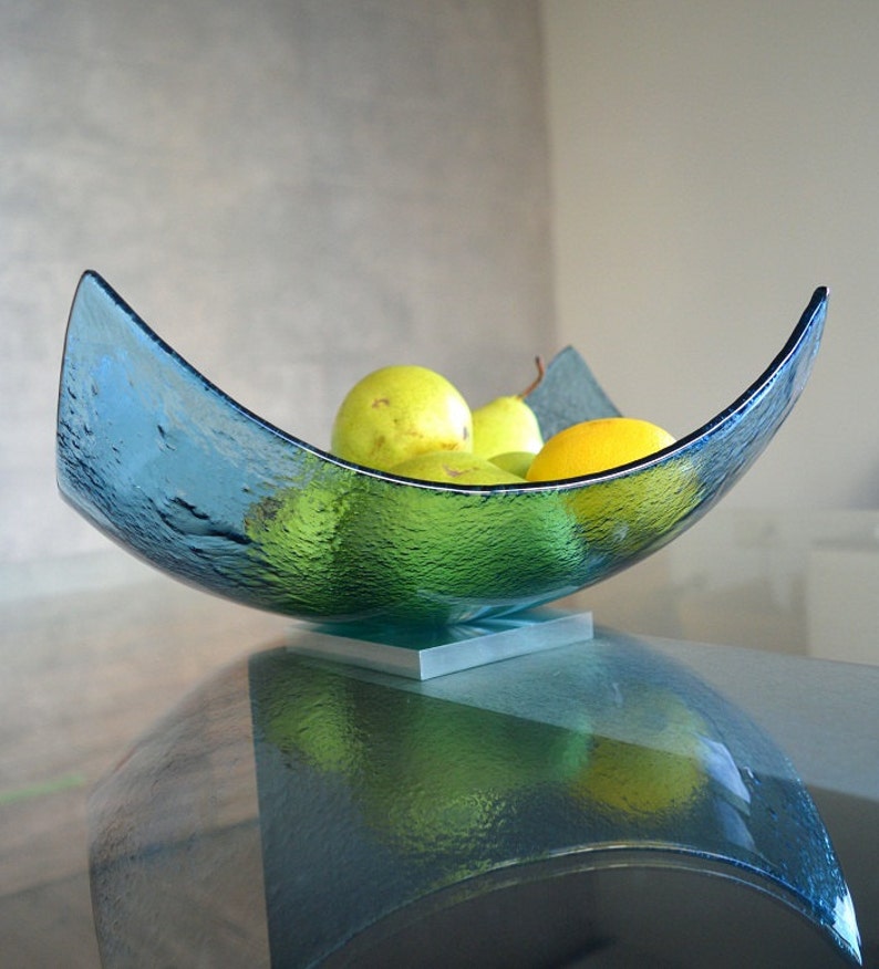 May include: A textured, turquoise glass bowl in a crescent shape, resting on a square metal base. The bowl is filled with green pears and a yellow lemon. The bowl is sitting on a glass table.