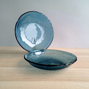 May include: Two blue glass bowls with textured surfaces and dark rims. One bowl is slightly elevated, showcasing the interior and rim detail. The bowls are on a light wood surface, with a neutral background.