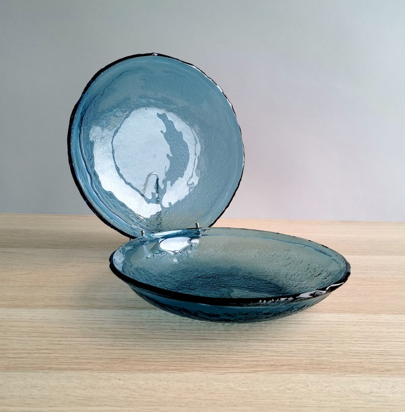May include: Two blue glass bowls with textured surfaces and dark rims. One bowl is slightly elevated, showcasing the interior and rim detail. The bowls are on a light wood surface, with a neutral background.
