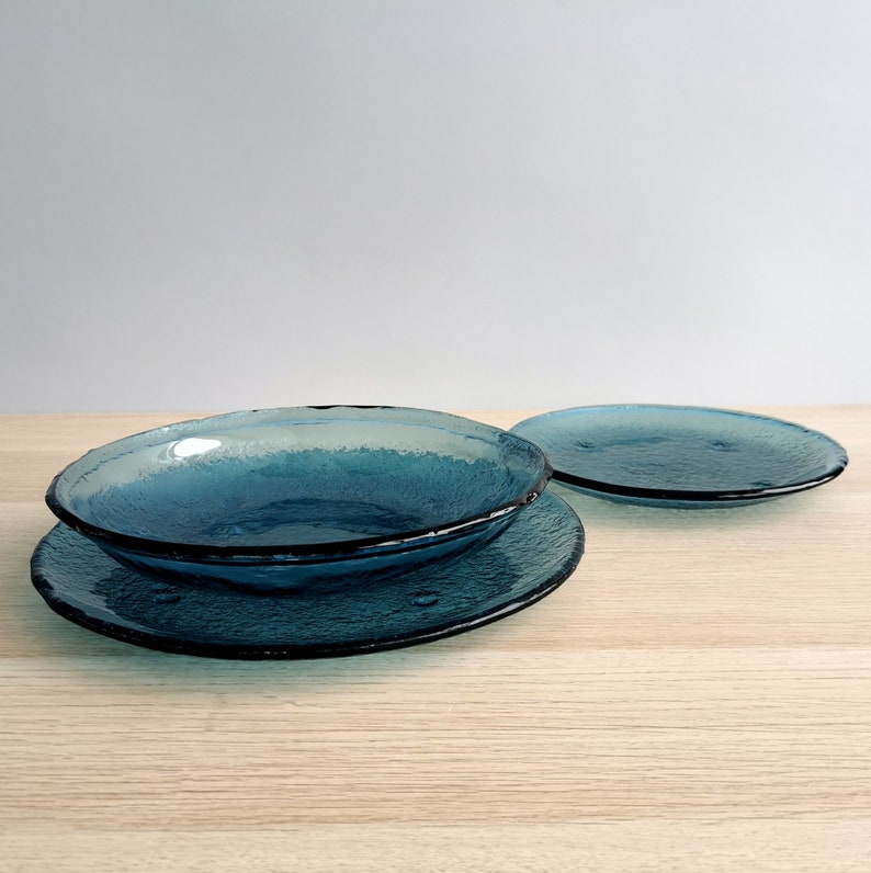 May include: A set of four textured, teal-colored glass dishes. The set includes two plates and two bowls. The plates and bowls have a dark teal rim. The dishes are arranged on a light wood surface.