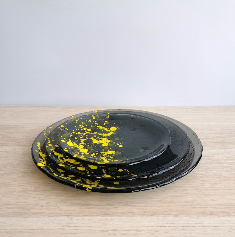 May include: Three black ceramic plates with yellow paint splatters. The plates are stacked on top of each other.