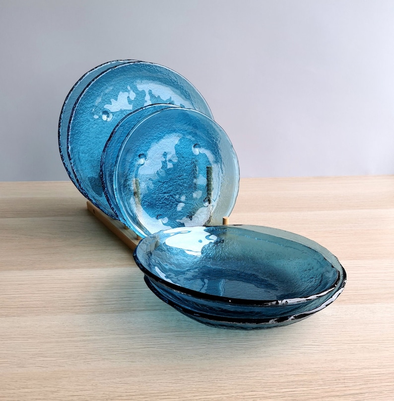 May include: A set of textured, turquoise-colored glass plates and bowls. The plates are displayed on a wooden rack, with a textured surface and a dark rim. The bowls are stacked in front.