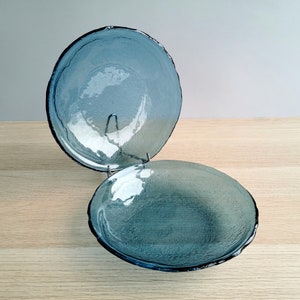 May include: Two blue glass bowls with textured surfaces and dark rims are displayed on a metal stand. The bowls have a round shape and a slightly irregular edge. The bowls are on a light wood surface.