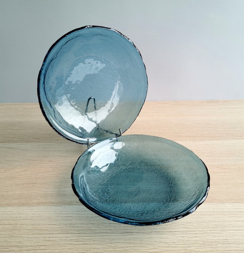 May include: Two blue glass bowls with textured surfaces and dark rims are displayed on a metal stand. The bowls have a round shape and a slightly irregular edge. The bowls are on a light wood surface.