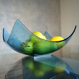 May include: A textured, teal-colored glass bowl with a unique, curved design. The bowl is filled with yellow lemons and green limes. It sits on a small, rectangular, white base. The bowl is displayed on a reflective surface.
