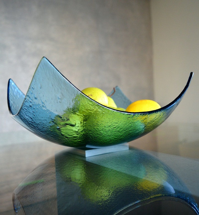 May include: A textured, teal-colored glass bowl with a unique, curved design. The bowl is filled with yellow lemons and green limes. It sits on a small, rectangular, white base. The bowl is displayed on a reflective surface.
