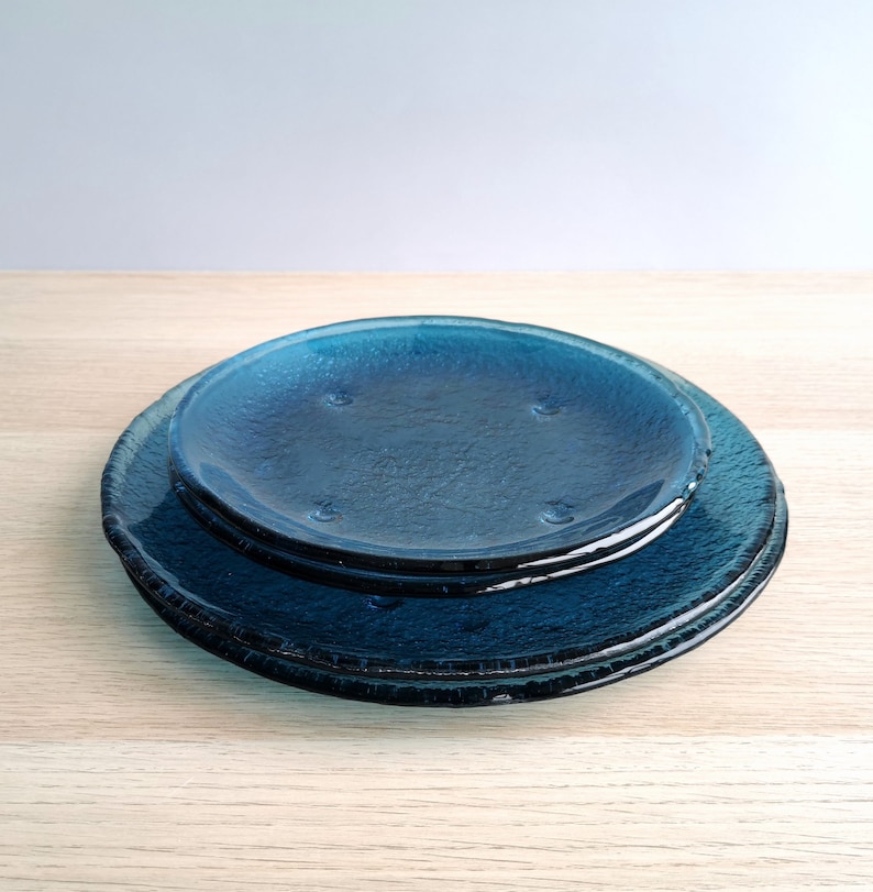 May include: A stack of three textured, round, blue glass plates. The plates have a slightly rough surface and a deep blue color. The plates are of different sizes, with the smallest plate on top.