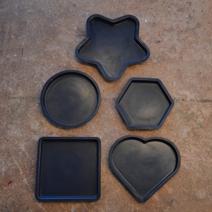 May include: Five black coasters in various shapes: a star, a circle, a hexagon, a square, and a heart. The coasters are made of a dark material and are arranged on a brown surface.
