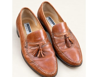 Könnte beinhalten: Ein Paar lohfarbene Leder-Slipper mit Quastendetail auf dem Vorderblatt und einer mokassinartigen Naht an der Schuhspitze. Das Innenfutter ist hell und enthält ein weißes Etikett mit dem schwarzen Aufdruck Florsheim Imperial.