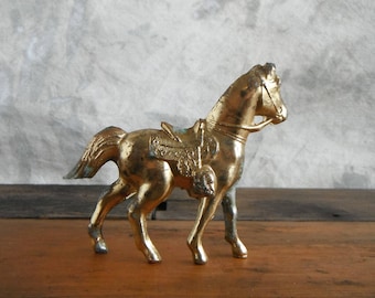 May include: A gold-colored metal horse figurine with a saddle and bridle. The horse is standing on a wooden surface.