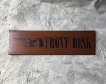 May include: A brown wooden sign with black text that reads "FRONT DESK". A black and white illustration of a hand pointing to the right is on the left side of the sign.