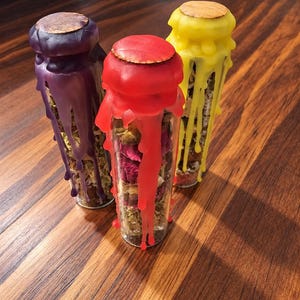 May include: Three clear glass cylindrical vials filled with dried herbs and flower petals. Each vial is sealed with a flat wooden stopper and topped with colored wax that drips down the sides in purple, red, and yellow hues.
