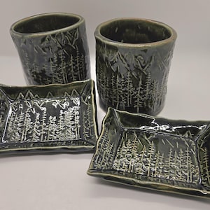 May include: A set of four ceramic pieces in a dark green glaze. Two cylindrical cups and two rectangular trays feature a carved mountain and forest design. The pottery has a rustic, handmade appearance, suitable for home decor or serving.