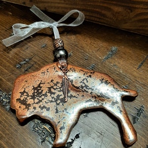 May include: A copper-colored buffalo ornament with a distressed finish. The ornament has a small feather charm hanging from it and a white ribbon for hanging.