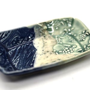 May include: A rectangular ceramic dish with a divided design. One side features a dark blue, swirling pattern, while the other has a light green, textured surface with a botanical motif. The dish is approximately 6 inches long.
