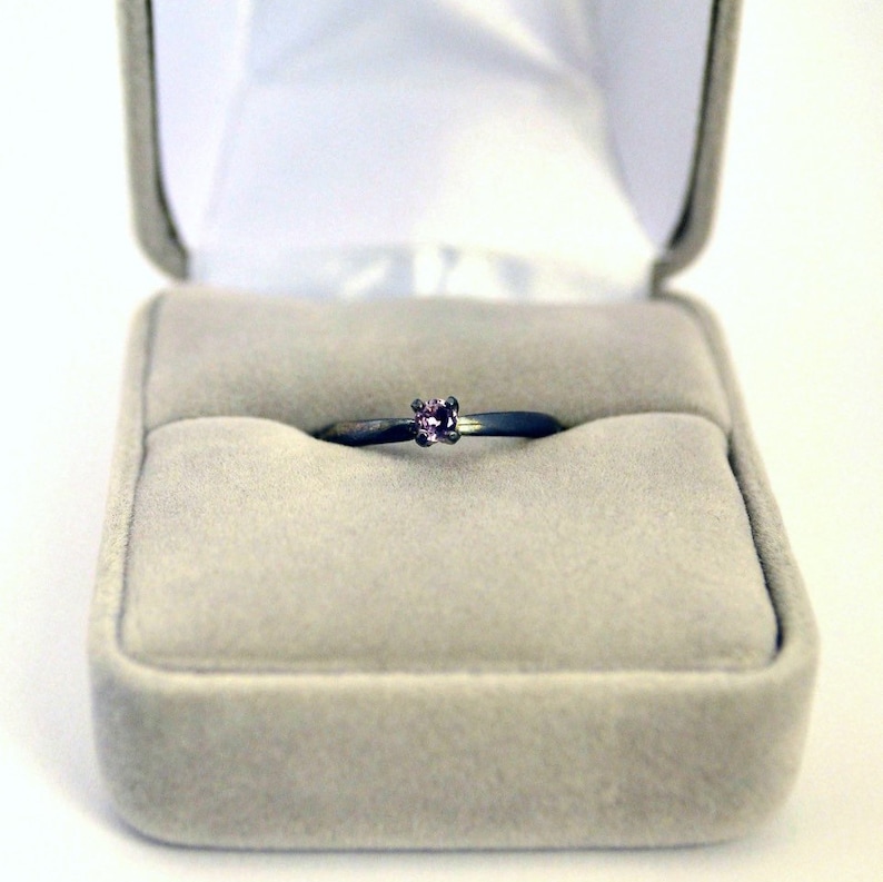 May include: A simple silver ring with a small pink gemstone set in the center. The ring is displayed in a gray velvet ring box.