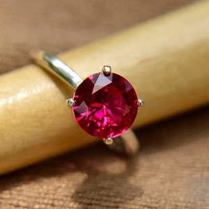 May include: A silver ring with a large, round, deep red ruby gemstone set in a four-prong setting. The ruby is 8 mm in diameter.