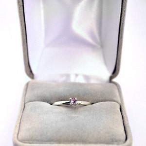 May include: A simple silver ring with a small, light pink gemstone set in the center, displayed in a gray velvet ring box.