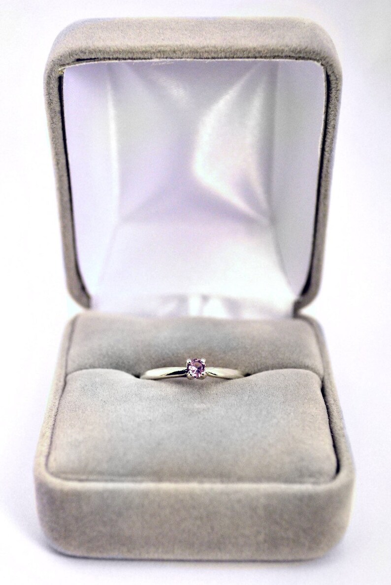 May include: A simple silver ring with a small, light pink gemstone set in the center, displayed in a gray velvet ring box.