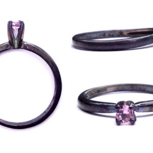 May include: A silver ring with a small pink gemstone set in a prong setting. The ring is oxidized for a darker finish.