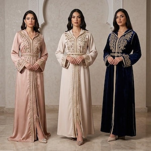 May include: Three floor-length kaftans featuring long sleeves, hoods, and intricate gold embroidery along the front placket, cuffs, and waist. The garments are presented in solid shades of dusty rose, light beige, and deep navy blue.