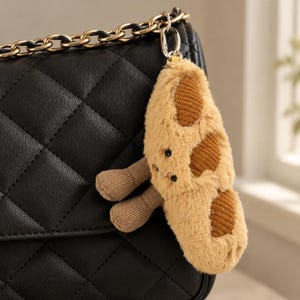 May include: A plush cookie-shaped bag charm with a fuzzy tan exterior, dark brown textured spots, and two small dangling legs. The charm features two black embroidered eyes and is attached to a gold-toned metal chain on a black quilted bag.