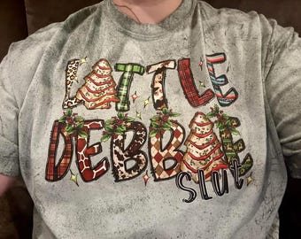 May include: A gray acid-wash t-shirt featuring the text "LITTLE DEBBIE SLUT" in large, multi-patterned letters. The design includes illustrations of Christmas tree cakes, holly sprigs, and small star accents in red, green, and brown.